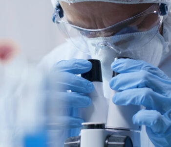 Closeup of scientist doctor looking into medical microscope analyzing blood sample during scientific experiment in biochemistry laboratory. Researcher man developing vaccine against covid19