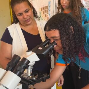 Estudante observa microscópio enquanto professora orienta (Foto/ Arquivo Pessoal).