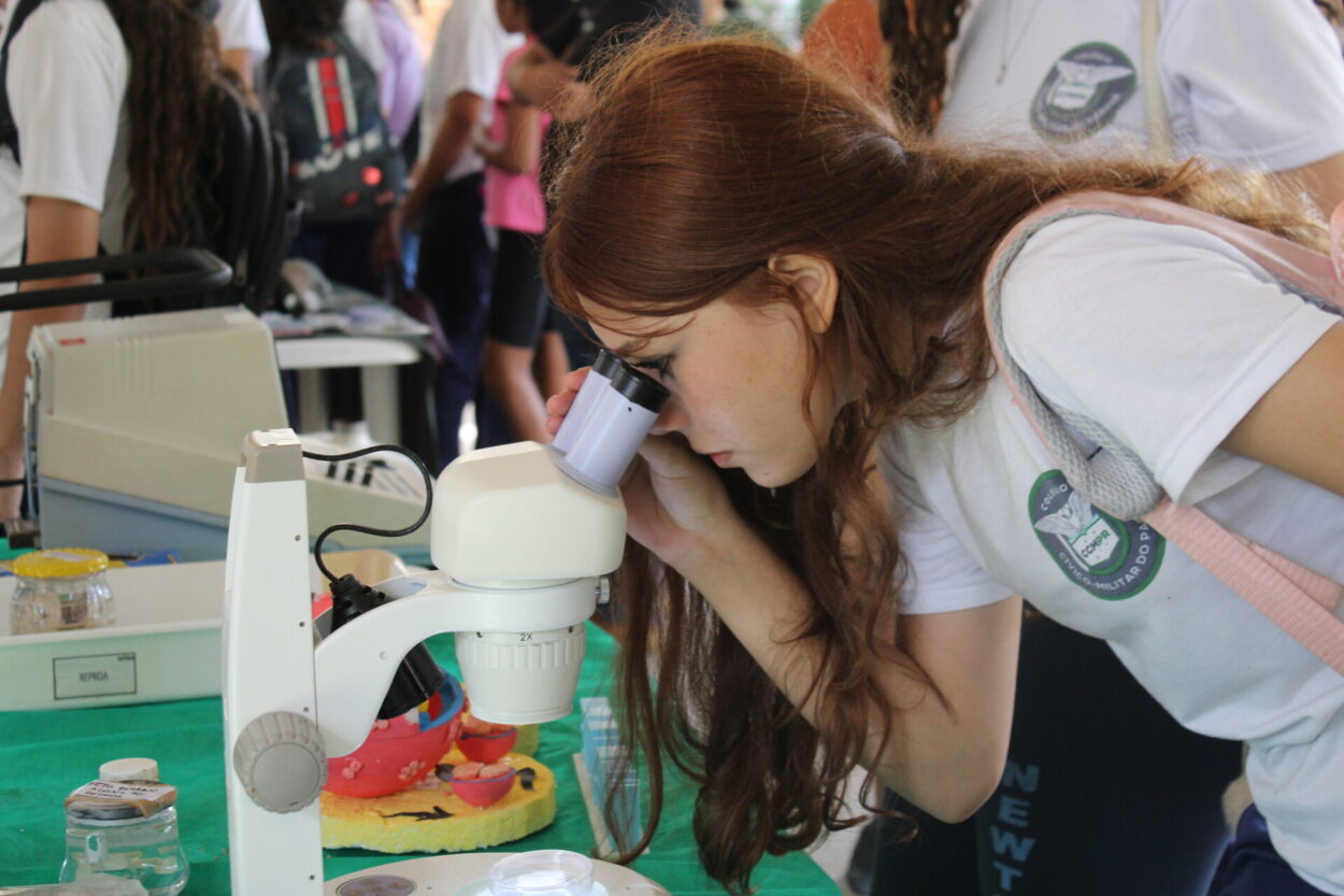 A imagem mostra uma estudante do clube Litterae Ludus visitando um dos estandes da Via Rural Smart Farm, na ExpoLondrina 2025. Uniformizada, a aluna participa de uma atividade proposta pelos monitores do estande e olha por um microscópio. No entorno, há outros alunos e pessoas com mochilas e na mesa, além do microscópio, há modelos didáticos coloridos (como uma maquete anatômica), recipientes com líquidos e outros materiais de laboratório.