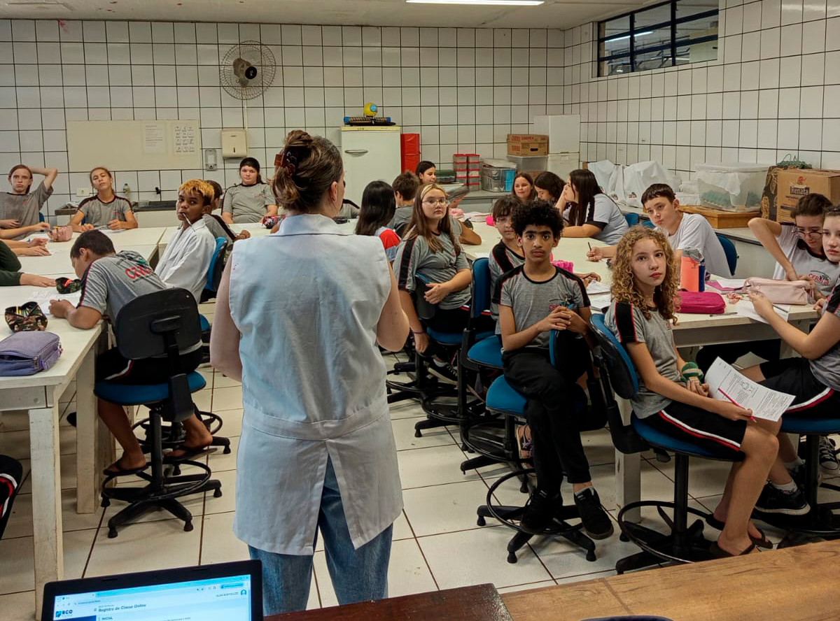 A imagem mostra uma sala de aula equipada como um laboratório, com alunos sentados em cadeiras giratórias ao redor de mesas brancas. A professora, vestida com um jaleco branco, está de costas para a câmera, aparentemente explicando algo para a turma. Os alunos, vestindo uniformes escolares cinza com detalhes vermelhos e pretos, exibem diferentes expressões e posturas – alguns atentos à professora, outros conversando ou olhando para os materiais em suas mesas. Alguns seguram folhas de papel e estojo de materiais escolares. O ambiente tem paredes revestidas com azulejos brancos, prateleiras com caixas e materiais organizados ao fundo, e um ventilador fixado na parede. No primeiro plano, um notebook está aberto, exibindo uma tela com um sistema online. O cenário sugere uma aula prática em um laboratório escolar.