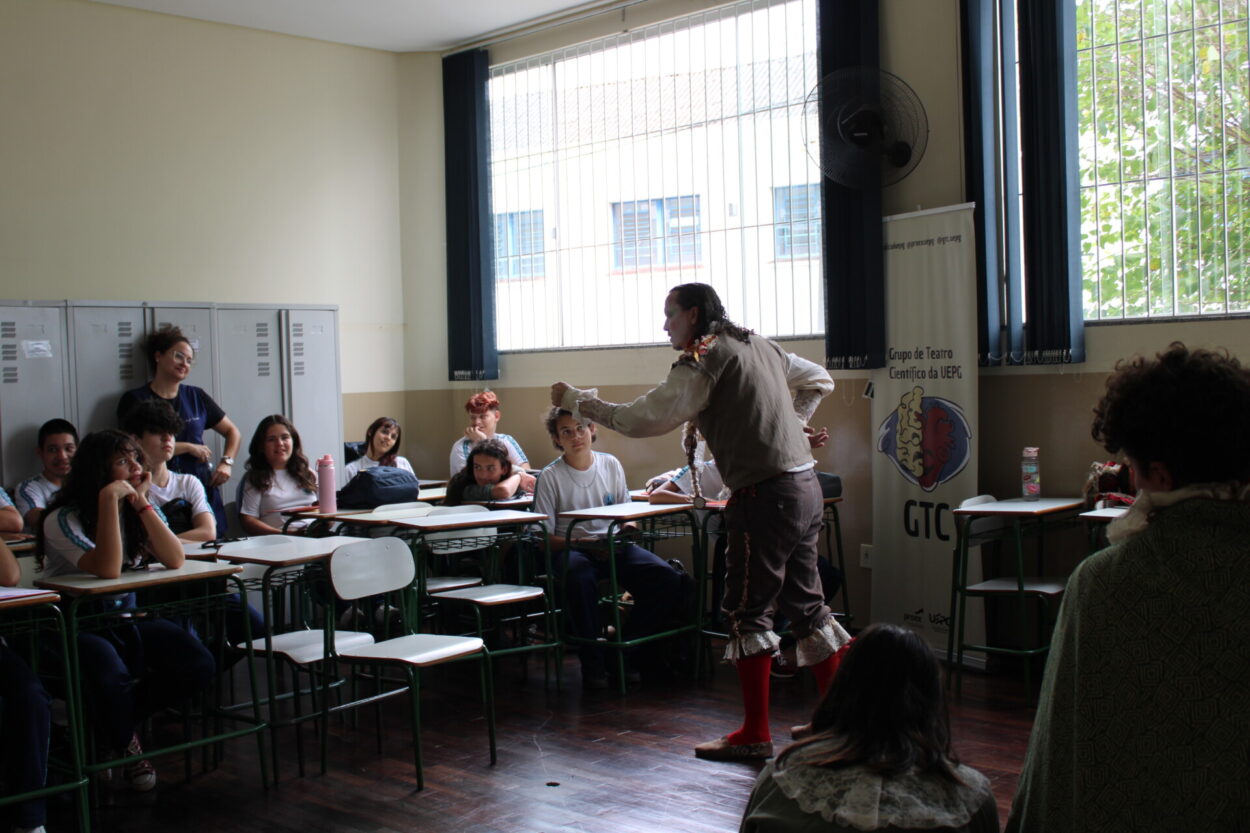 A imagem retrata uma cena teatral em sala de aula, onde um ator caracterizado com vestimentas de época — incluindo meias vermelhas, colete e babados — apresenta-se diante de estudantes sentados, atentos e sorridentes. Ao fundo, há lockers escolares, janelas com persianas verticais abertas deixando entrar luz natural, e um banner com o logo do Grupo de Teatro Científico da UEPG (GTC). A interação entre o artista e os alunos parece criar uma atmosfera de envolvimento, sugerindo uma ação educativa que une arte, ciência e ensino de forma lúdica e reflexiva.