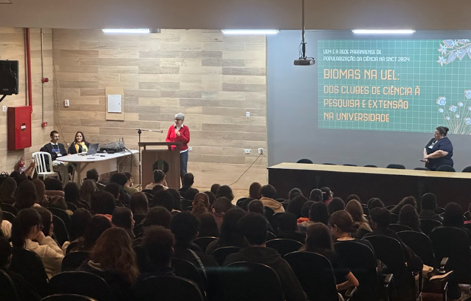 A imagem mostra um auditório com estudantes assistindo à palestra de abertura do evento realizado na UEL. Uma mulher com blusa vermelha está em pé falando ao microfone. À esquerda, duas pessoas estão sentadas acompanhando a apresentação. No canto direito do palco, uma mulher está observando a fala. No telão, está projetado o título do evento: "Biomas na UEL: dos clubes de ciência à pesquisa e extensão na universidade", com o subtítulo: "UEM e a rede paranaense de popularização da ciência na SNCT 2024". O ambiente é fechado, com iluminação artificial, paredes revestidas com paineis de madeira clara e cadeiras pretas.
