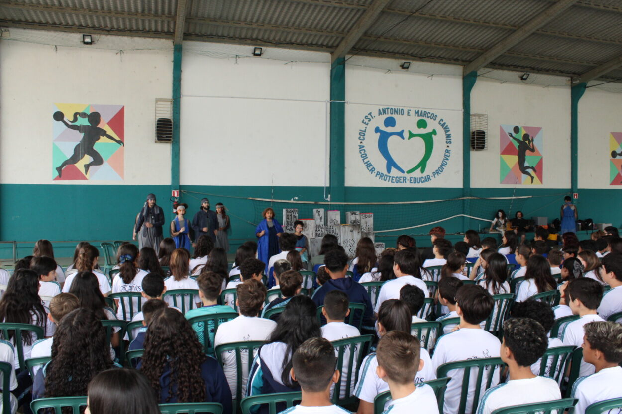 A imagem mostra uma apresentação teatral em andamento em um ginásio escolar coberto, diante de um grande grupo de estudantes sentados em cadeiras plásticas verdes, todos vestindo uniformes brancos com detalhes azuis. No palco improvisado, seis pessoas caracterizadas com figurinos em tons de cinza e azul atuam diante de um cenário que simula um muro pichado com palavras e símbolos. Ao fundo, nas paredes, há murais coloridos com silhuetas esportivas e o logotipo da escola “Col. Est. Antonio e Marcos Cavanis” com o lema “Acolher, Proteger, Educar, Promover”. À direita, duas pessoas operam o som próximo a uma mesa com equipamentos. A cena transmite um ambiente escolar durante atividade cultural