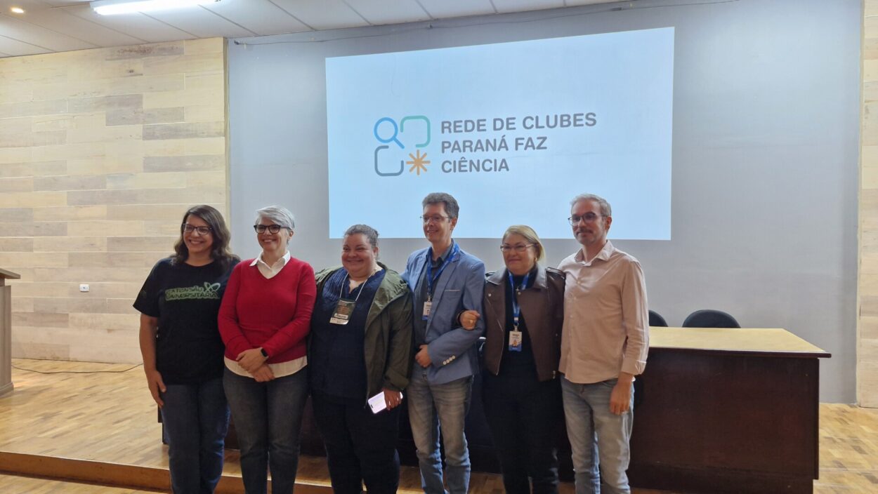 Na imagem, seis pessoas estão lado a lado no palco, posando para a foto em um auditório. Ao fundo, o telão exibe o logotipo da Rede de Clubes Paraná Faz Ciência. A foto foi tirada em um ambiente com piso de madeira e paredes claras. Elas são palestrantes de um evento do NAPI "Paraná Faz Ciência", realizado na Universidade Estadual de Londrina. As pessoas estão vestidas com roupas casuais e formais.