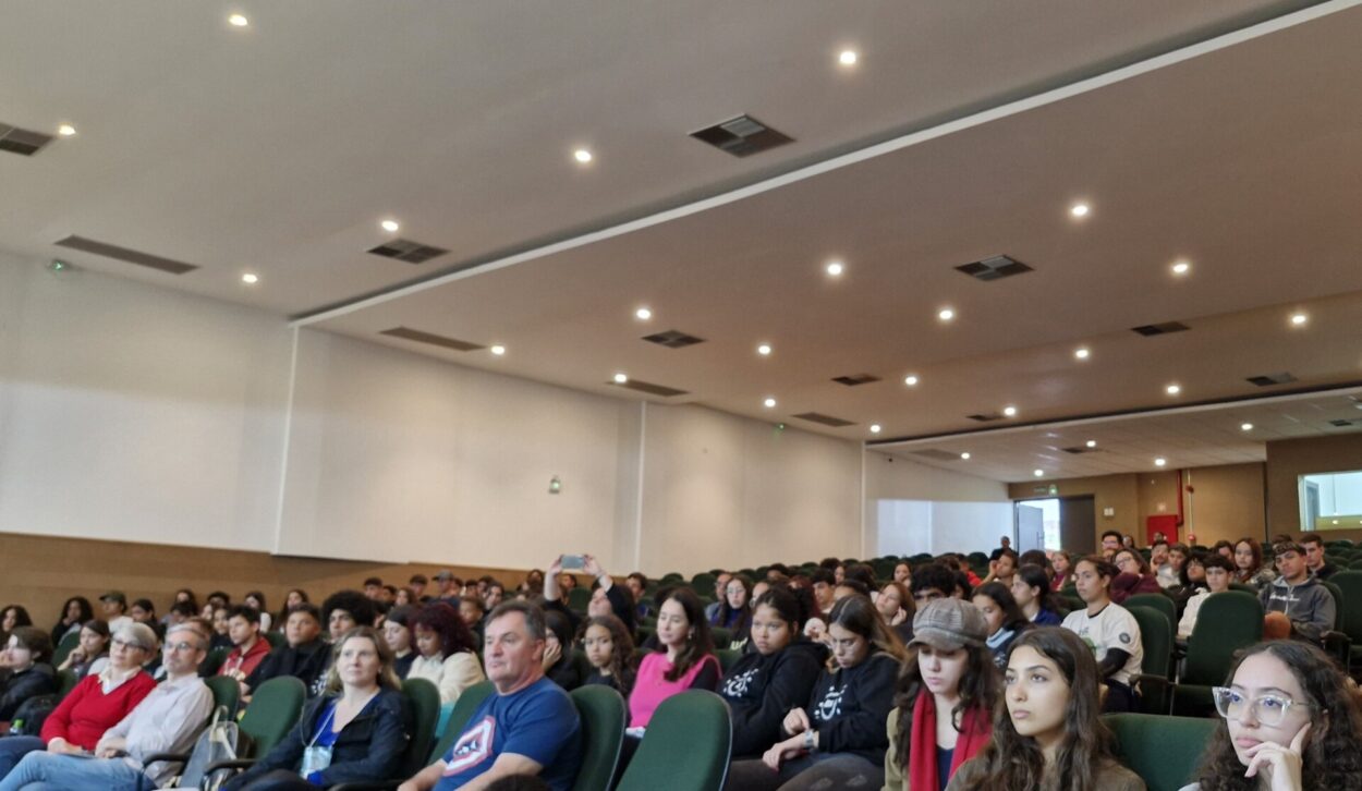 A imagem mostra um auditório lotado, com estudantes e professores prestando a atenção no que estava sendo apresentado. O espaço é amplo, com poltronas verdes e iluminação de teto com luzes embutidas, proporcionando um ambiente bem iluminado. A cena indica que trata-se de um evento acadêmico.