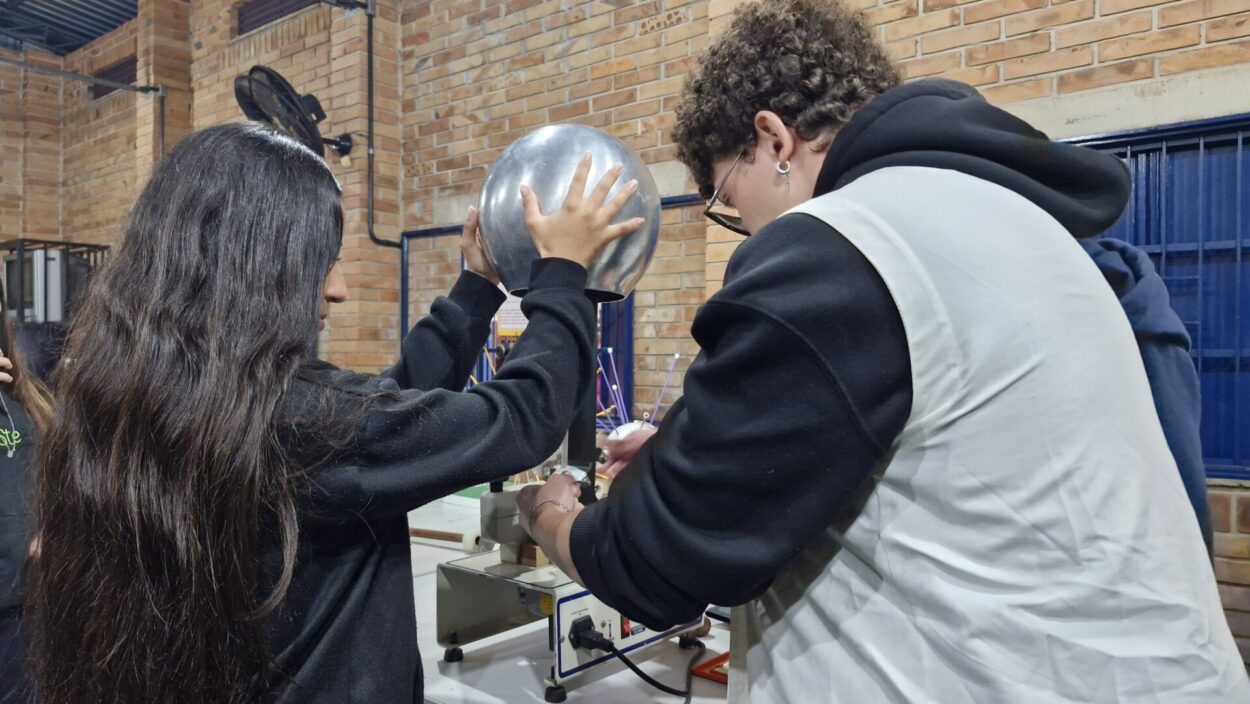 A imagem mostra uma estudante uniformizada participando de um experimento sobre eletrização com um gerador de Van de Graaff. No centro da imagem, a aluna com cabelos longos e escuros está de perfil, tocando com as duas mãos uma grande esfera metálica na parte superior do gerador. Ao lado dela, um pesquisador manuseia o equipamento. A atividade ocorre em um ambiente fechado com paredes de tijolos à vista e janelas com grades.