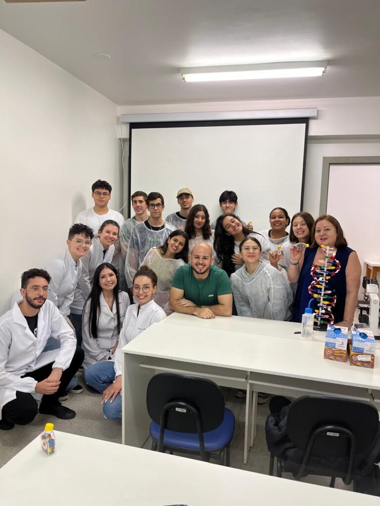A imagem mostra um grupo de pessoas reunidas no Laboratório de Imunogenética. Entre eles, há estudantes, professores e profissionais vestidos com jalecos brancos. Todos estão sorrindo e posando para a foto, com um homem de camiseta verde ao centro apoiado em uma mesa. No ambiente, há uma tela de projeção ao fundo, microscópio, caixas de leite e um modelo de DNA colorido sobre a mesa, indicando atividades ligadas à ciência e biologia. A atmosfera parece descontraída e colaborativa, sugerindo um evento educacional.