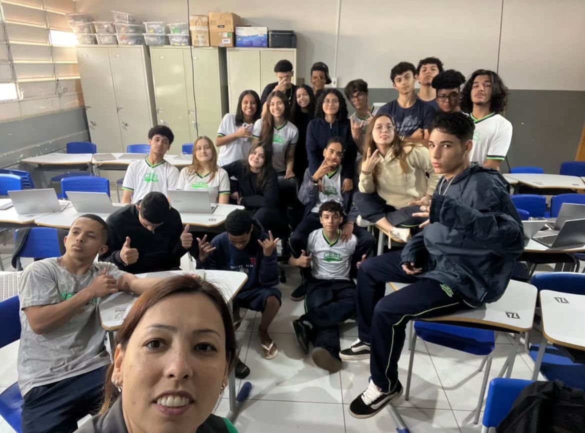 A imagem foi fotografada no primeiro dia do Clube de Ciências Capivara´s Tech no ano passado. Podemos ver um grupo de estudantes em uma sala de aula, posando para uma selfie tirada por uma mulher na frente da câmera. A turma é composta por jovens com diferentes estilos e expressões, eles vestem uniformes escolares brancos com detalhes verdes. A sala tem carteiras organizadas, algumas com laptops sobre elas, e armários no fundo. A iluminação natural entra pelas janelas laterais. Os estudantes parecem descontraídos, com alguns fazendo gestos com as mãos e outros sorrindo ou mantendo expressões mais sérias. O ambiente reflete um clima de interação entre os alunos e a professora.