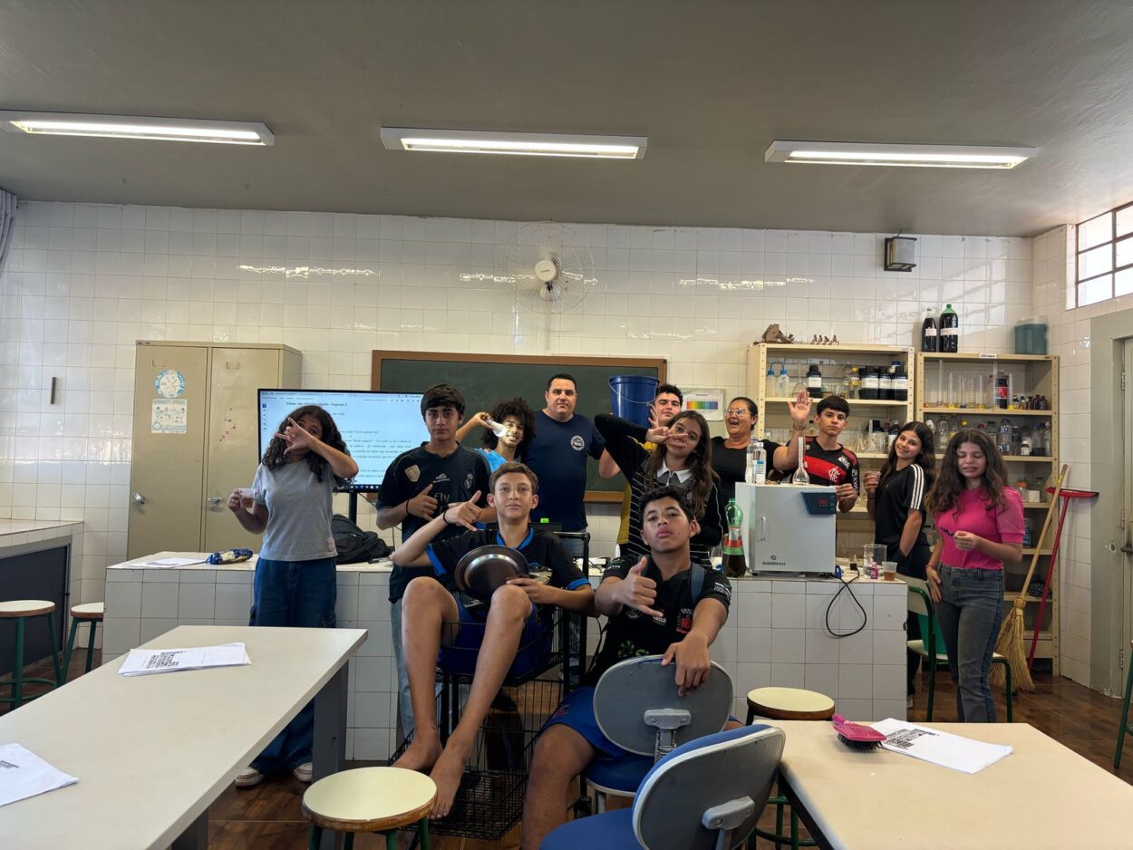 A foto é do Clube de Ciências Show da Química. A imagem mostra um grupo de estudantes e um professor em um laboratório de química. O ambiente possui prateleiras com vidrarias e reagentes químicos, além de uma lousa e um monitor ao fundo exibindo um texto. Os alunos estão interagindo de forma descontraída, fazendo gestos e poses variadas. Alguns seguram objetos como uma panela e uma garrafa, e há materiais sobre as mesas, sugerindo que estão realizando uma atividade experimental. O clima parece de descontração e colaboração.