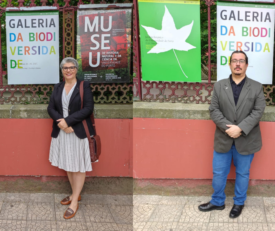 A imagem é uma montagem composta por duas fotos lado a lado, tiradas em frente a painéis informativos do Museu de História Natural e da Ciência da Universidade do Porto, mais especificamente da Galeria da Biodiversidade e do Jardim Botânico da Universidade do Porto. Foto à esquerda: uma mulher está em pé, sorridente, usando um vestido listrado em tons claros, blazer preto e sapatos marrons. Ela carrega uma bolsa de ombro marrom e usa óculos. Está posicionada em frente a um cartaz que diz "GALERIA DA BIODIVERSIDADE". Foto à direita: um homem está em pé, com expressão séria, vestindo uma calça jeans azul, blazer cinza, camisa preta e sapatos pretos. Ele está em frente a outro cartaz da "GALERIA DA BIODIVERSIDADE" e ao lado de um painel verde com uma folha branca estilizada, associado ao Jardim Botânico da universidade. Ambas as pessoas parecem estar visitando o local em caráter institucional, dado o traje formal-casual e o cenário acadêmico.