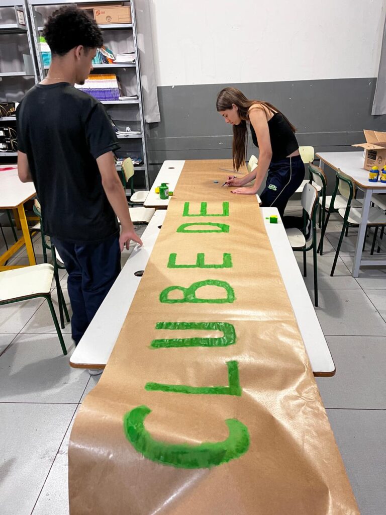 A imagem mostra dois jovens trabalhando na confecção de um cartaz em uma sala de aula. No centro da imagem, há uma faixa longa de papel kraft estendida sobre duas mesas escolares, unidas para formar uma superfície contínua. Na faixa, está sendo pintada a frase "CLUBE DE" com letras grandes e verdes. À direita, uma jovem com cabelos longos e castanhos está ajoelhada sobre uma cadeira enquanto pinta as letras. À esquerda, um jovem observa o trabalho. Ao fundo, há estantes metálicas com livros, pastas e caixas organizadoras. O ambiente tem aparência escolar, com paredes cinza e branca, cadeiras escolares e materiais de arte sobre as mesas. A cena retrata um momento de colaboração e criatividade.