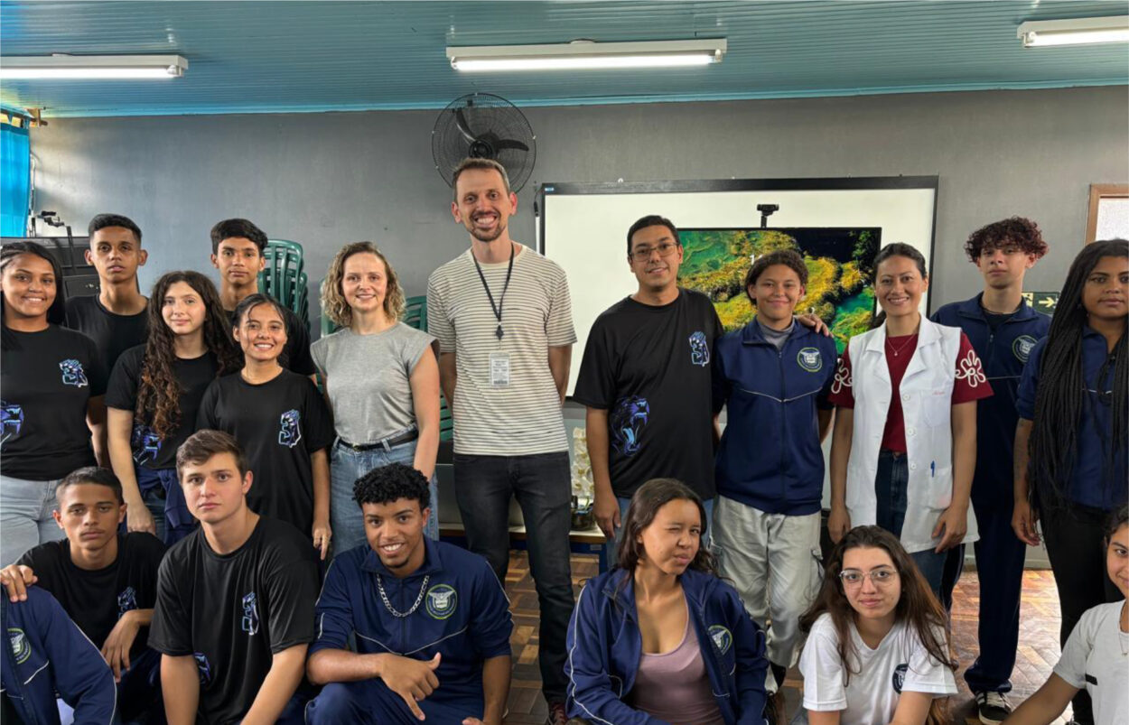 A imagem mostra um grupo de pessoas em uma sala de aula. O grupo é composto por estudantes e professores, alguns vestindo uniformes escolares e outros com roupas casuais. Há uma tela ao fundo exibindo uma imagem da natureza, possivelmente um ambiente subaquático ou florestal. O ambiente tem um teto azul com luminárias e ventiladores. Algumas pessoas estão sorrindo, enquanto outras mantêm expressões neutras.