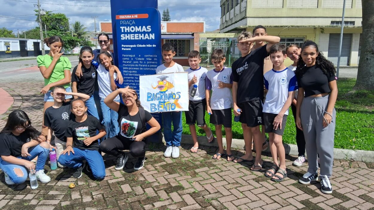 Um grupo de 16 pessoas, a maioria crianças e adolescentes, está reunido em uma praça ao lado de uma placa azul que indica o nome do local: "Praça Thomas Sheehan". A placa menciona que a praça é uma homenagem a um paraquedista canadense herói de guerra. O grupo segura um cartaz colorido com os dizeres "Bagrinhos do Bento" e um desenho de um peixe. Algumas crianças estão fazendo poses divertidas, cobrindo os olhos com as mãos para proteger-se do sol. A maioria veste roupas casuais, como camisetas e calças jeans, e alguns estão calçando chinelos ou tênis. Ao fundo, há construções, árvores e uma calçada de tijolos intertravados.