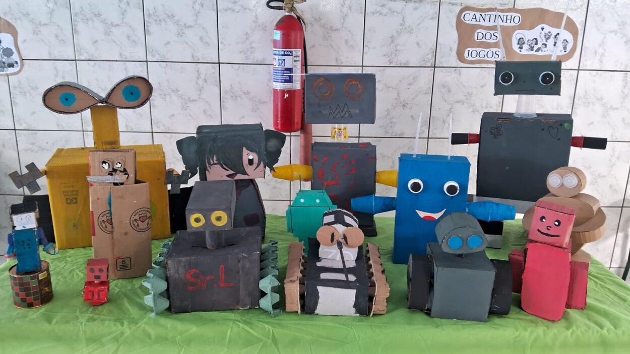 Foto de uma mesa coberta com um tecido verde, sobre a qual estão expostos vários robôs coloridos feitos com materiais recicláveis como caixas de papelão, latas e rolos de papel. Cada robô tem um formato diferente e muitos apresentam olhos desenhados ou colados com tampinhas. Ao fundo, há uma parede com azulejos brancos e um cartaz escrito "Cantinho dos Jogos". Os robôs variam em tamanho e expressão, compondo uma mostra criativa de trabalhos escolares.