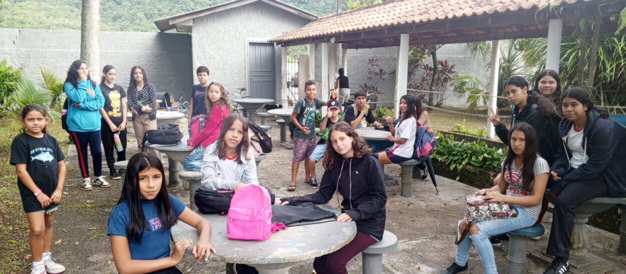 Foto de um grupo de aproximadamente 20 estudantes reunidos em um pátio externo arborizado, com mesas e bancos de concreto. Alguns alunos estão sentados, outros em pé, com mochilas e bolsas ao lado. Ao fundo, há um muro cinza, uma edificação com telhado de telha vermelha e vegetação densa com montanhas verdes ao longe. A maioria dos estudantes usa roupas casuais e há expressões diversas — alguns sorrindo, outros mais sérios, olhando para a câmera.