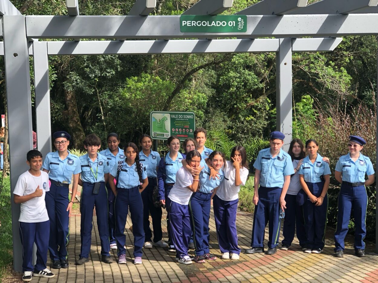 Na imagem, um grupo de jovens, alguns vestidos com uniformes azuis com boinas e outros de camisetas brancas, posando em frente a uma estrutura de madeira com a placa "PERGOLADO 01". Atrás deles, há uma placa verde com informações sobre o local, que parece ser uma área de preservação ambiental, com árvores e vegetação ao fundo. O grupo está sorrindo e fazendo poses variadas, demonstrando um momento de confraternização ao ar livre.