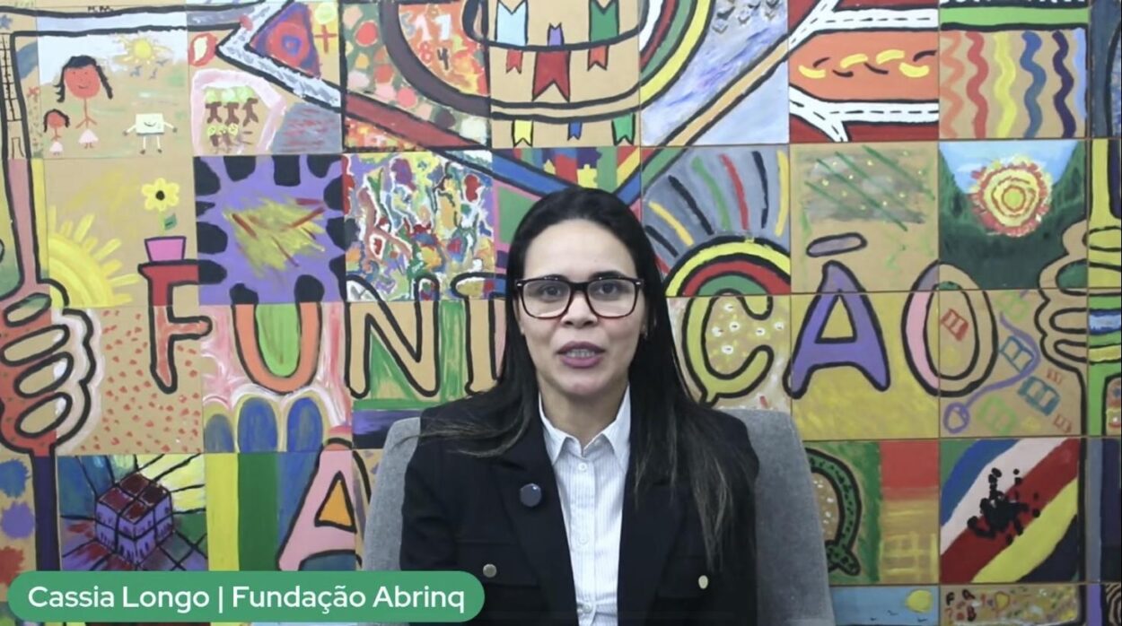 Na captura de tela da live de lançamento do Projeto Árvore, uma mulher de pele clara, cabelos lisos e escuros, usando óculos de armação preta, blazer preto e camisa branca, está sentada em uma cadeira cinza. Ao fundo, há um mural colorido com desenhos infantis e a palavra “FUNDAÇÃO” pintada em letras grandes e vibrantes. No canto inferior da imagem, uma faixa verde com o texto: “Cassia Longo | Fundação Abrinq”.