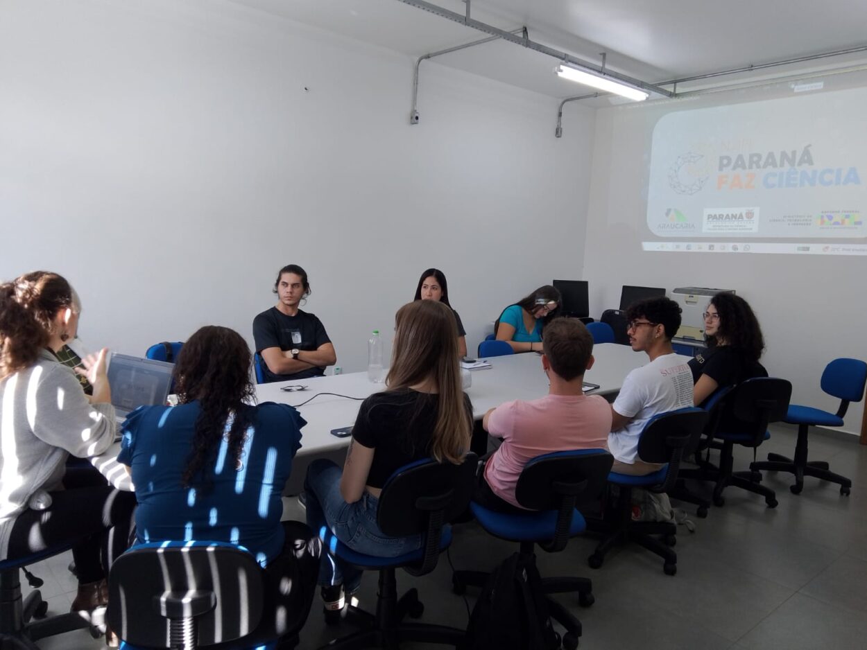 A foto mostra a equipe da UEPG reunida ao redor de uma mesa em uma sala. Uma apresentação do projeto "Paraná Faz Ciência" é projetada na parede ao fundo. Uma pessoa, posicionada à esquerda da imagem, utiliza um notebook enquanto conversa com o grupo. Outras pessoas acompanham a fala, com anotações e materiais sobre a mesa. O espaço conta com iluminação natural e artificial.