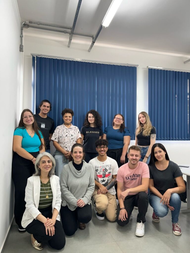 A foto mostra o grupo organizado para uma foto coletiva em uma sala, com cortinas azuis ao fundo. Algumas pessoas estão em pé e outras estão agachadas ou ajoelhadas à frente. Todos olham para a câmera.