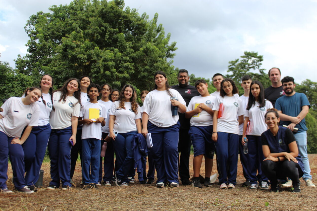 A imagem mostra os estudantes e professores que fazem parte do clube “Discovery Kennedy’s”, do Colégio Estadual Cívico-Militar Presidente Kennedy, de Rolândia. A turma realizou uma visita à Fazenda Bimini, na zona rural da cidade. A foto é um registro do passeio. Os alunos estão uniformizados e em volta há apenas vegetação.