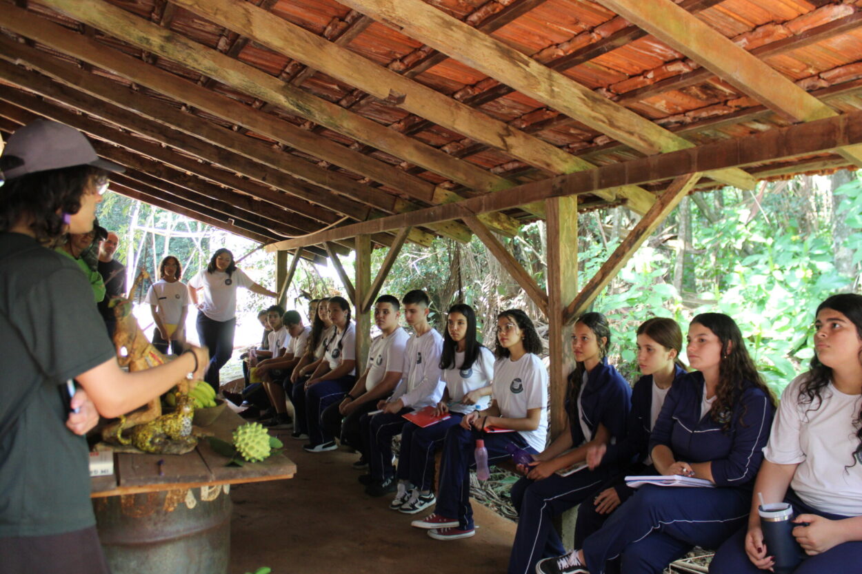 A imagem mostra os estudantes do clube “Discovery Kennedy’s”, do Colégio Estadual Cívico-Militar Presidente Kennedy, de Rolândia, prestando atenção na aula sobre natureza e simbiose. A turma realizou uma visita à Fazenda Bimini, na zona rural da cidade, e receberam explicações sobre biodiversidade e floresta nativa. A foto é um registro do passeio. Os alunos estão uniformizados, sentados em um banco de madeira. Na frente deles, tem uma mesa com objetivos e um estagiário de biologia, que acompanhou o passeio e estava se apresentando.