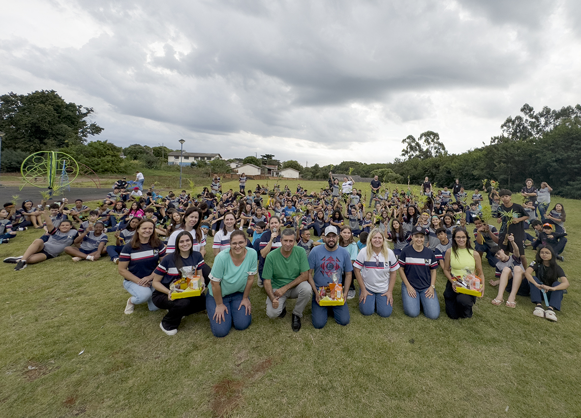 A imagem mostra um grande grupo de pessoas reunidas ao ar livre, em um campo gramado, com muitas árvores ao fundo sob um céu nublado. Na parte da frente da imagem, há uma fileira de adultos (professores e palestrantes) agachados e atrás deles, há uma grande quantidade de crianças e adolescentes sentados ou em pé, muitos segurando pequenas mudas de plantas. No canto esquerdo da imagem, é possível ver parte de um parquinho infantil com brinquedos, e ao fundo algumas casas e postes de iluminação. Todos parecem estar felizes, sorrindo para a foto.