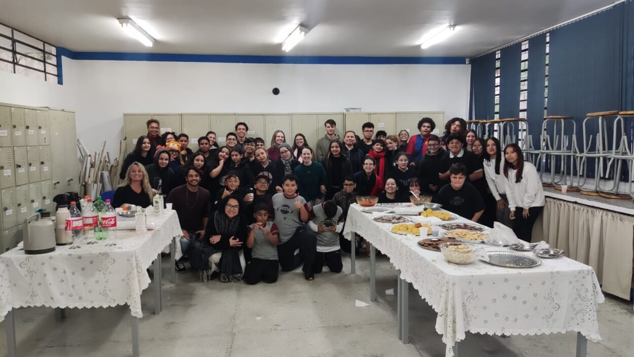 A imagem mostra um grupo grande de pessoas reunidas em uma sala de uma escola, participando de confraternização. O ambiente é fechado, com luzes fluorescentes no teto, armários ao fundo e várias janelas com cortinas azul-escuras do lado direito. O grupo, composto por crianças, adolescentes e alguns adultos, está reunido ao redor de duas mesas cobertas com toalhas brancas rendadas. As mesas estão repletas de alimentos e bebidas, como bolos, salgados, refrigerantes e sucos. Todos estão sorrindo ou posando para a foto, criando um clima alegre e descontraído.