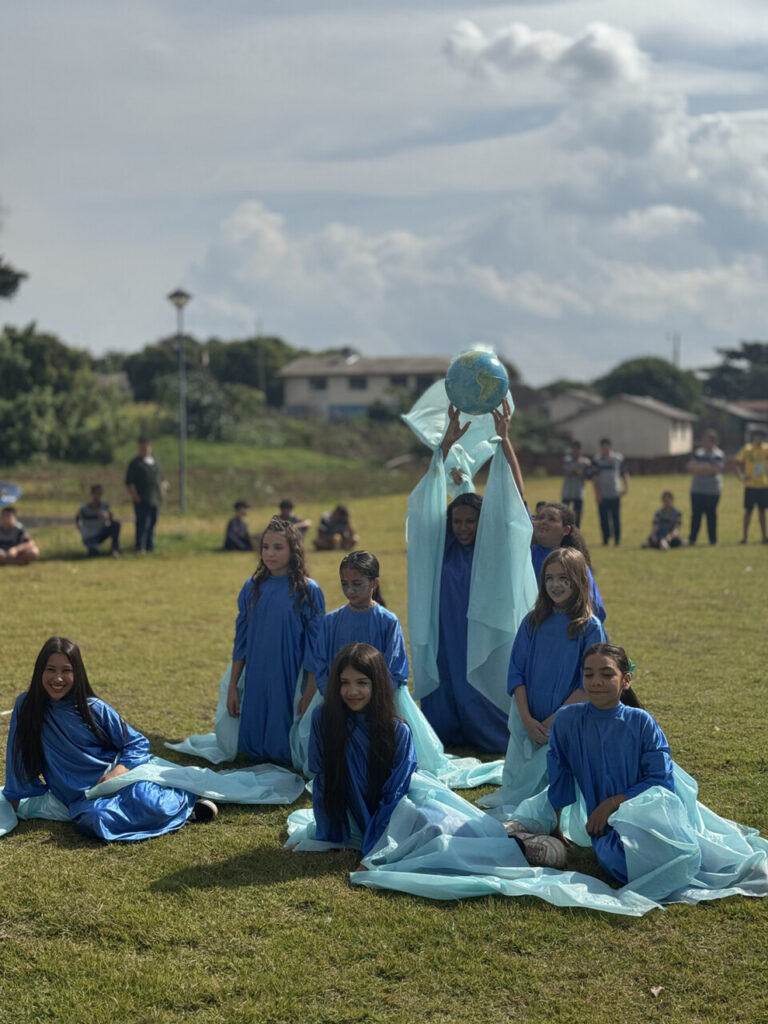 A imagem mostra um grupo de meninas participando de uma apresentação ao ar livre, elas são membros do clube de dança da escola. Elas estão vestidas com túnicas em tons de azul e azul-claro, remetendo à água. Uma das meninas, posicionada ao centro e em pé, segura um globo terrestre acima da cabeça, o que reforça a temática ambiental e de conscientização ecológica. As meninas estão organizadas em uma composição coreografada, com expressões alegres e concentradas. Algumas têm o rosto pintado como elemento artístico ou teatral. Ao fundo, há pessoas observando a apresentação, algumas sentadas na grama, outras em pé. O céu está parcialmente nublado, com uma luz suave incidindo sobre o grupo, criando um efeito bonito na cena. A imagem transmite uma sensação de harmonia, cuidado com o planeta e valorização da expressão artística na escola.