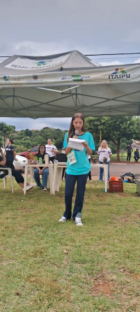 A imagem mostra uma menina em pé, em um espaço ao ar livre, lendo um texto em voz alta com a ajuda de um microfone. Ela está segurando algumas folhas de papel e parece estar concentrada na leitura. A menina veste camiseta azul e calça escura, com um crachá pendurado no pescoço. Ao fundo, há uma tenda com logotipos de instituições como IDR-Paraná, Embrapa, Itaipu Binacional e o brasão do município de Borrazópolis, indicando a parceria entre diferentes órgãos em um evento educacional e ambiental. Atrás dela, algumas pessoas estão sentadas e observando atentamente, entre elas adultos e estudantes. Também é possível ver mesas plásticas com vasos de plantas, reforçando o tema ambiental, e ao fundo, carros e árvores, indicando que o evento ocorre em uma área aberta urbana, próxima a escola. A cena transmite a ideia de um momento importante de participação estudantil pelo meio ambiente, educação e cidadania.