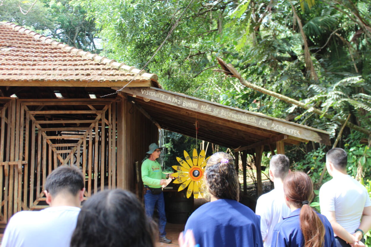 A imagem mostra o ambientalista Daniel Steidle apresentando a Fazenda Bimini aos estudantes do clube “Discovery Kennedy’s”, do Colégio Estadual Cívico-Militar Presidente Kennedy, de Rolândia. Na foto, ao fundo, Daniel segura um girassol de madeira com os princípios da ONU escritos nas pétalas. Na frente, os alunos estão de costas prestando atenção na explicação e realizando anotações.