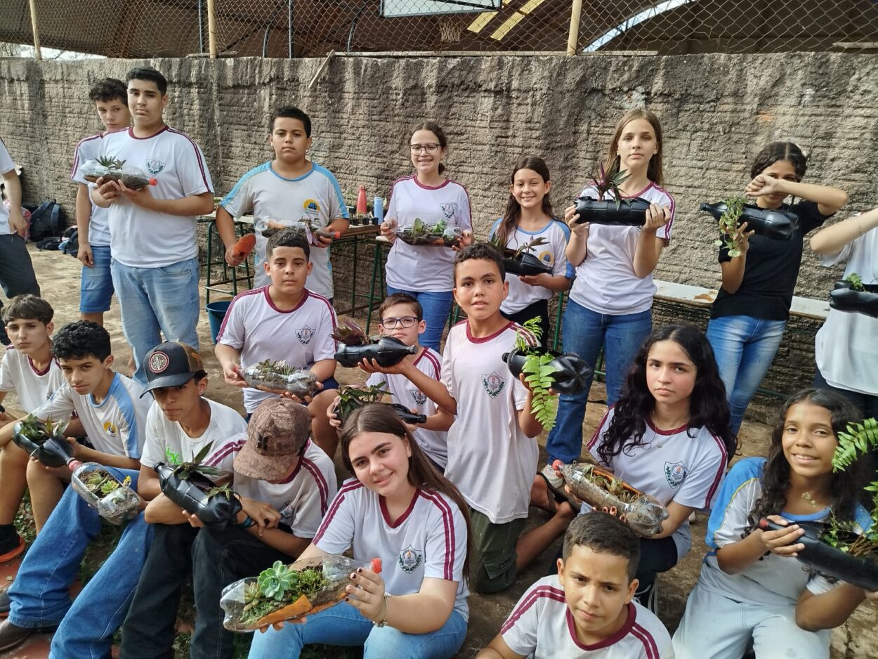 Esta imagem mostra um grupo de alunos envolvidos numa atividade prática com foco em reciclagem. Os estudantes estão a segurar vasos feitos a partir de garrafas plásticas reutilizadas, preenchidas com terra e várias plantas, como suculentas e pequenas folhagens. Os alunos estão organizados em duas filas, alguns agachados e outros de pé, e vestem uniformes escolares. O ambiente é ao ar livre, com uma parede de cimento ao fundo e uma rede de proteção no topo. A expressão dos alunos reflete orgulho e empenho no projeto.