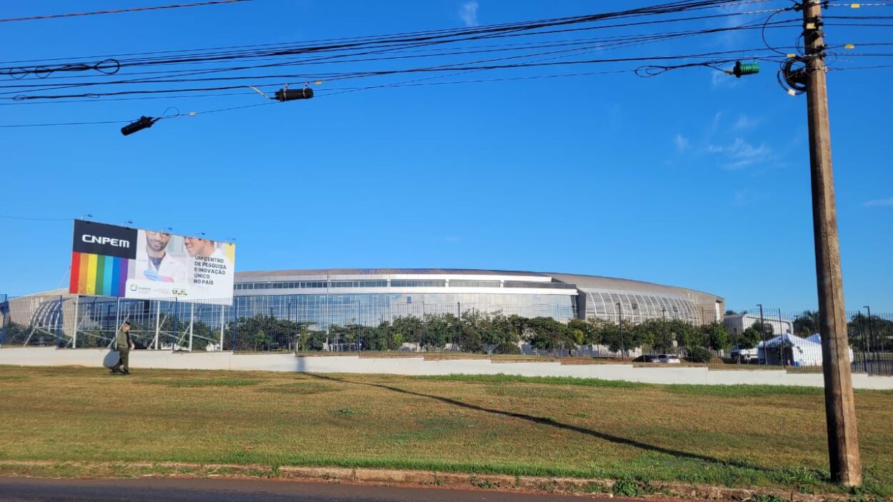 A imagem mostra o prédio visto de fora e de longe, em formato arredondado onde está o acelerador de partículas. A fachada é envidraçada, lembrando uma estrutura de estádio esportivo grandioso e ao lado esquerdo há uma placa estilo outdoor escrito: “CNPEM um centro de pesquisa e inovação único no país”. No entorno, a área é gramada e sem nenhuma construção em volta