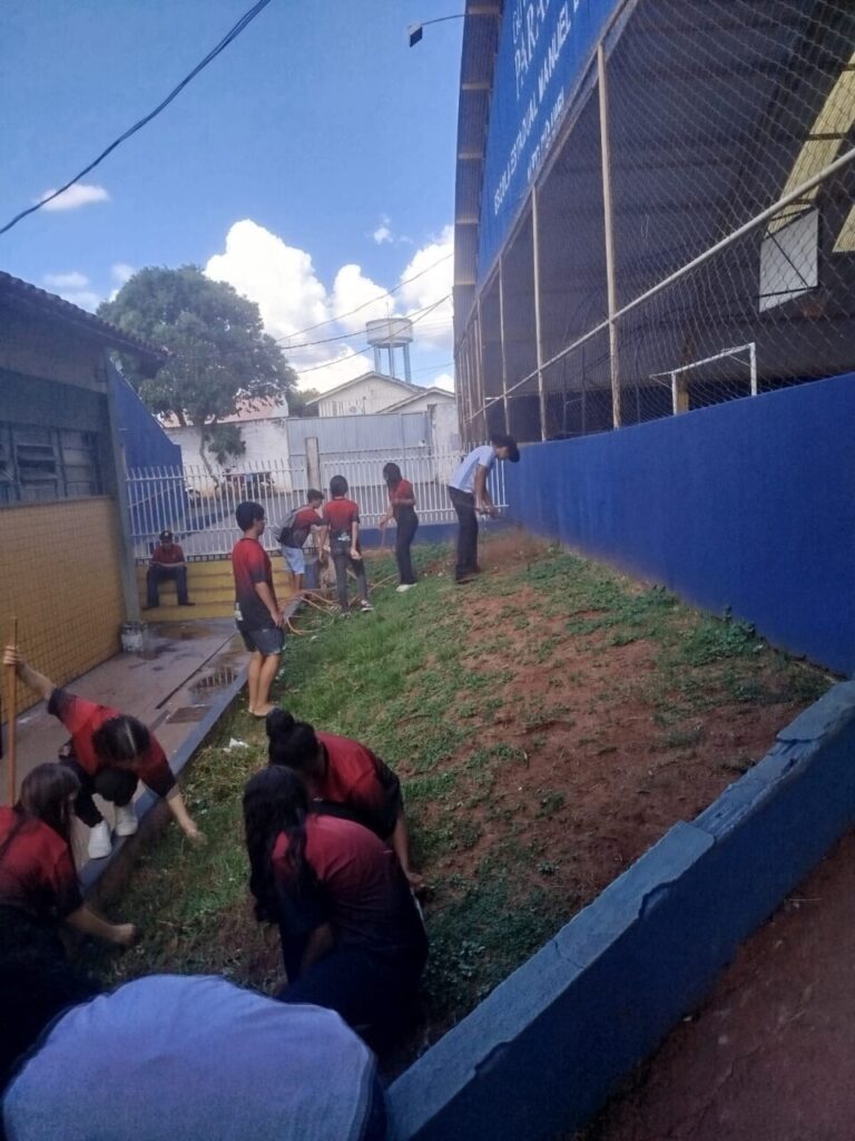 Esta imagem retrata um grupo de alunos a participar numa atividade de jardinagem. A ação decorre ao longo de um corredor externo com uma área de terra e relva junto a um muro azul, provavelmente lateral a uma quadra esportiva coberta (identificável pela rede e estrutura ao fundo). Os estudantes estão usando camisetas vermelhas do uniforme escolar. Alguns estão ajoelhados no chão, atentos ao trabalho manual, enquanto outros manuseiam ferramentas ou mangueiras para irrigação.
