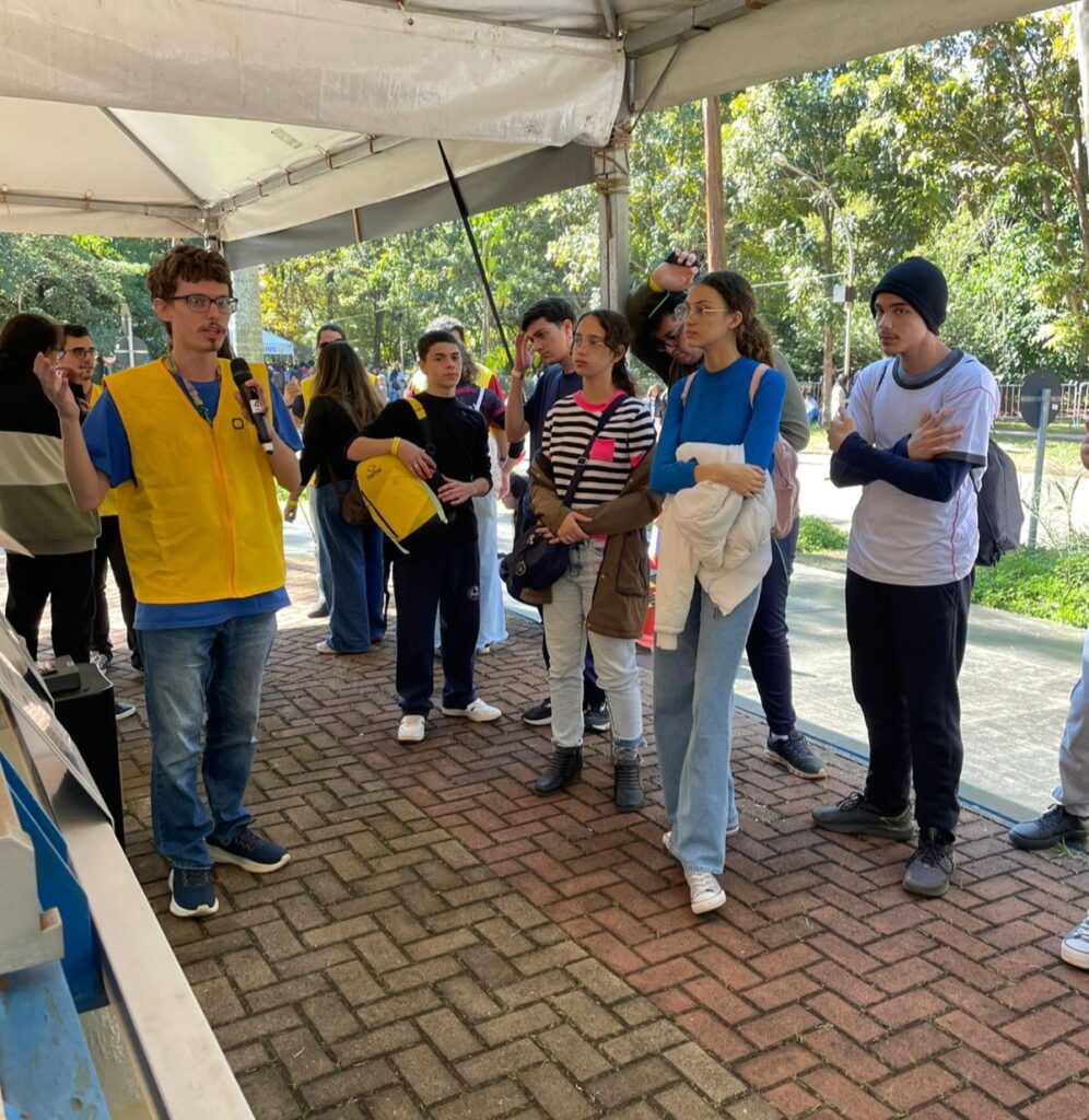 A imagem mostra um grupo de alunos em pé, em frente a um equipamento, e ao lado deles está um monitor que veste colete amarelo e segura um microfone para explicar o funcionamento do acelerador de partículas em miniatura. Estão numa área externa, porém embaixo de uma barraca ou estande montado pelo evento.