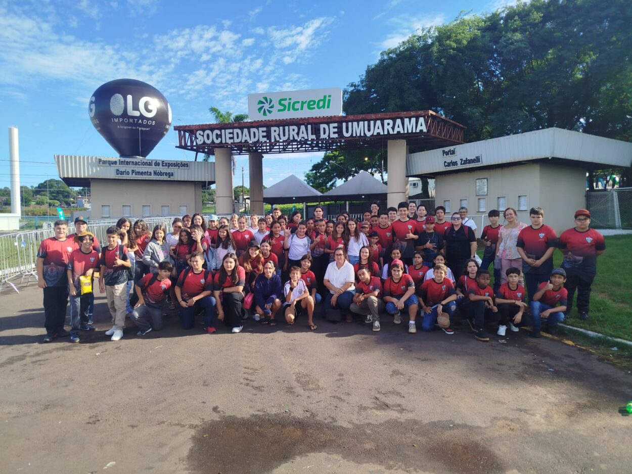 Esta imagem mostra um grande grupo de alunos e professores reunidos em frente à entrada do Parque Internacional de Exposições Dario Pimenta Nóbrega, localizado na Sociedade Rural de Umuarama. A maioria dos alunos está usando uniformes escolares com camisetas vermelhas e pretas, indicando que pertencem à mesma instituição. O grupo posa sorridente e animado.