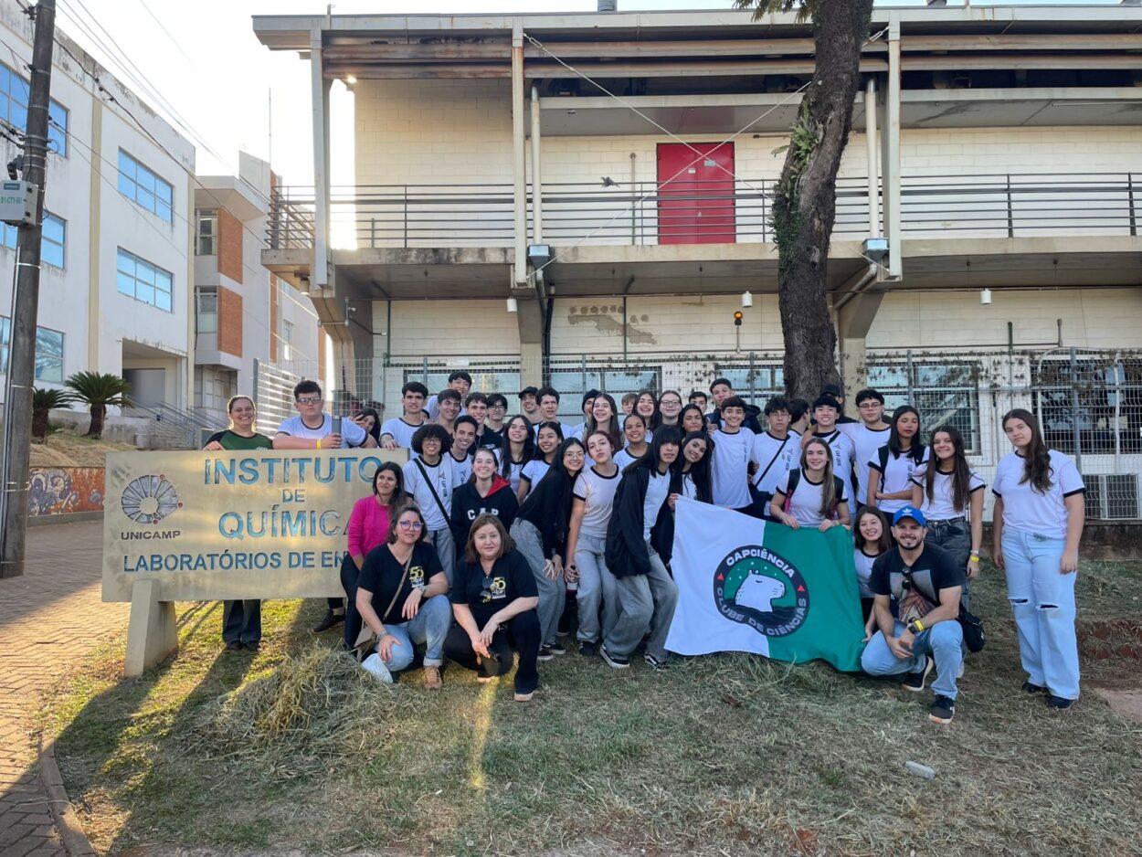 A imagem mostra um grupo de alunos e de professores alguns em pé, outros ajoelhados segurando a bandeira do Clube de Ciência CAPciência e posam ao lado da placa do Instituto de Química da UNICAMP (Universidade Estadual De Campinas). Ao fundo estão prédios do centro de estudos de química, no campus da UNICAMP.