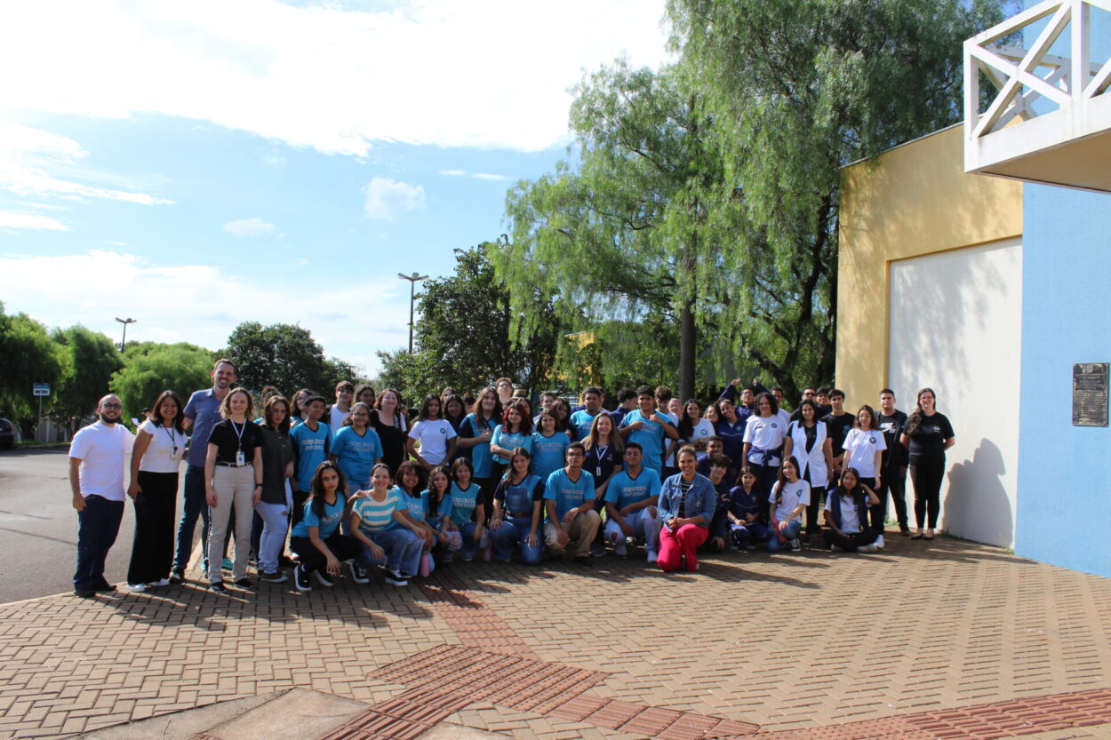 Foto de um grande grupo de cerca de 60 pessoas, entre jovens e adultos, reunidos ao ar livre em um espaço pavimentado, em frente a um prédio de paredes amarelas e azuis. Muitos usam camisetas azuis claras ou brancas, indicando participação em um evento conjunto. As pessoas estão sorrindo e posando para a foto, transmitindo um clima de alegria e união. Ao fundo, há árvores verdes e um céu azul com algumas nuvens.