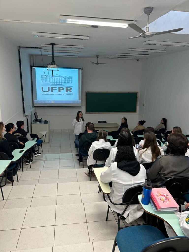 A galeria de fotos mostra momentos da visita dos clubistas à UFPR. Trata-se de ambientes fechados como um auditório e um laboratório. Há fotos da palestra que mostra várias cadeiras enfileiradas ocupadas por estudantes e professores, que estão assistindo à palestrante e à projeção de slide sobre o Rio iguaçu. Nas fotos do laboratório, mostra pequenos grupos de estudantes ouvindo orientações dos professores, usando microscópios e tablets, fazendo anotações. Todos estão vestindo jalecos com expressões de concentração e interesse.