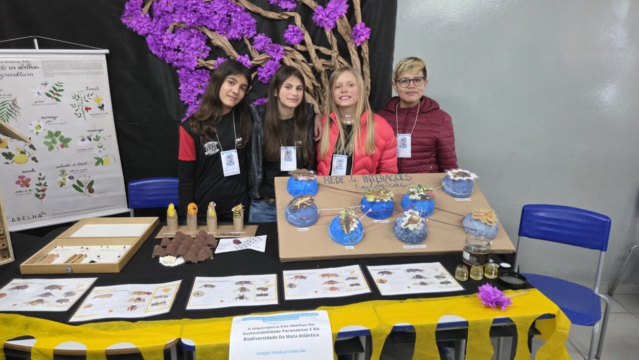 Quatro estudantes do Colégio Estadual Cristo Rei estão em pé atrás de uma mesa expositiva decorada com materiais didáticos sobre abelhas. São três meninas e uma menina. Eles usam crachás da Fieg e estão sorrindo. A mesa traz maquetes coloridas representando uma "rede de interações ecológicas", pequenos frascos com mel, cartazes informativos e modelos de abelhas. Ao fundo, há um mural com uma grande árvore feita de papel kraft com flores roxas em destaque. Um dos cartazes destaca a importância das abelhas na agricultura. A apresentação faz parte do projeto vencedor da Fieg na categoria Clubes de Ciências.