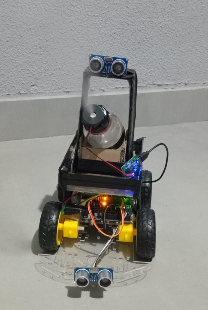 Esta imagem mostra um robô desenvolvido de forma artesanal. O robô possui uma estrutura montada com rodas, sensores ultrassônicos na parte frontal e superior, e componentes eletrônicos conectados, como placas de controle e fiação exposta. Na parte superior do robô, há um pequeno reservatório acoplado a um sistema que emite uma névoa, indicando a pulverização de um líquido, como parte da função do robô no combate à dengue. O sistema parece ter sido projetado para se locomover de maneira autônoma ou controlada, detectando obstáculos através dos sensores e liberando o produto quando necessário, visando eliminar ou repelir o mosquito Aedes aegypti. Esse tipo de projeto integra tecnologia e soluções sustentáveis para problemas reais, como o combate à dengue.