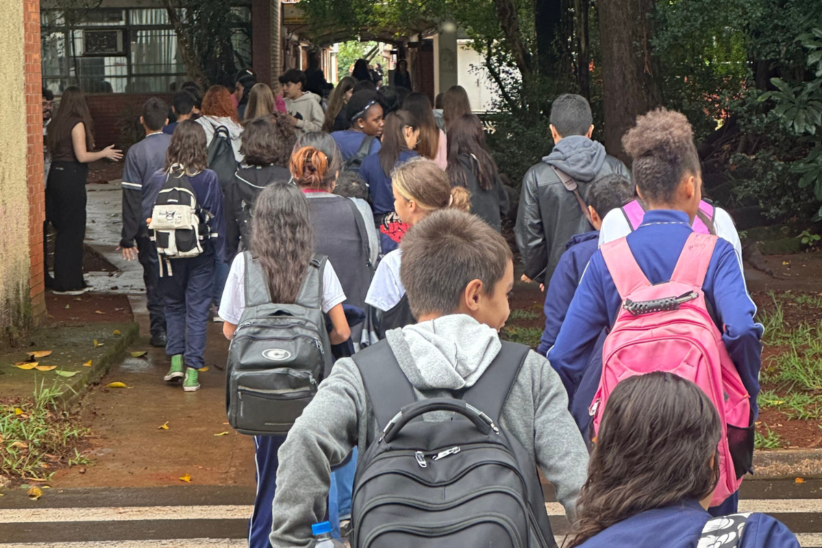 A imagem mostra vários alunos, com mochilas nas costas, saindo de um ônibus. Os estudantes estavam fazendo uma visita à Universidade Estadual de Londrina. Alguns alunos também estão uniformizados. Os adolescentes atravessam uma rua, para adentrar no Centro de Ciências Biológicas. No canto esquerdo, uma das bolsistas orienta os estudantes a seguirem o caminho certo.