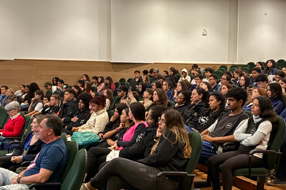 A imagem mostra vários estudantes e professores sentados em um anfiteatro da Universidade Estadual de Londrina (UEL). As pessoas estão concentradas, prestando a atenção no que estava sendo dito no palco. Alguns alunos estão uniformizados, enquanto outros foram com roupas do dia a dia. As cadeiras são verdes e as paredes claras.