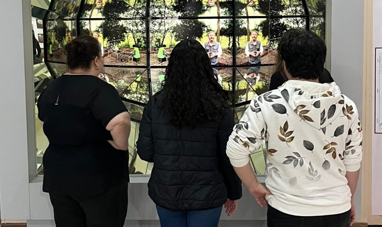 A imagem mostra uma das exposições do MUDI. Três pessoas estão de costas, observando um painel multimídia composto por várias telas espelhadas. No centro, há um vídeo educativo. As telas refletem e multiplicam as imagens, criando um efeito visual envolvente e imersivo que chama a atenção dos visitantes.