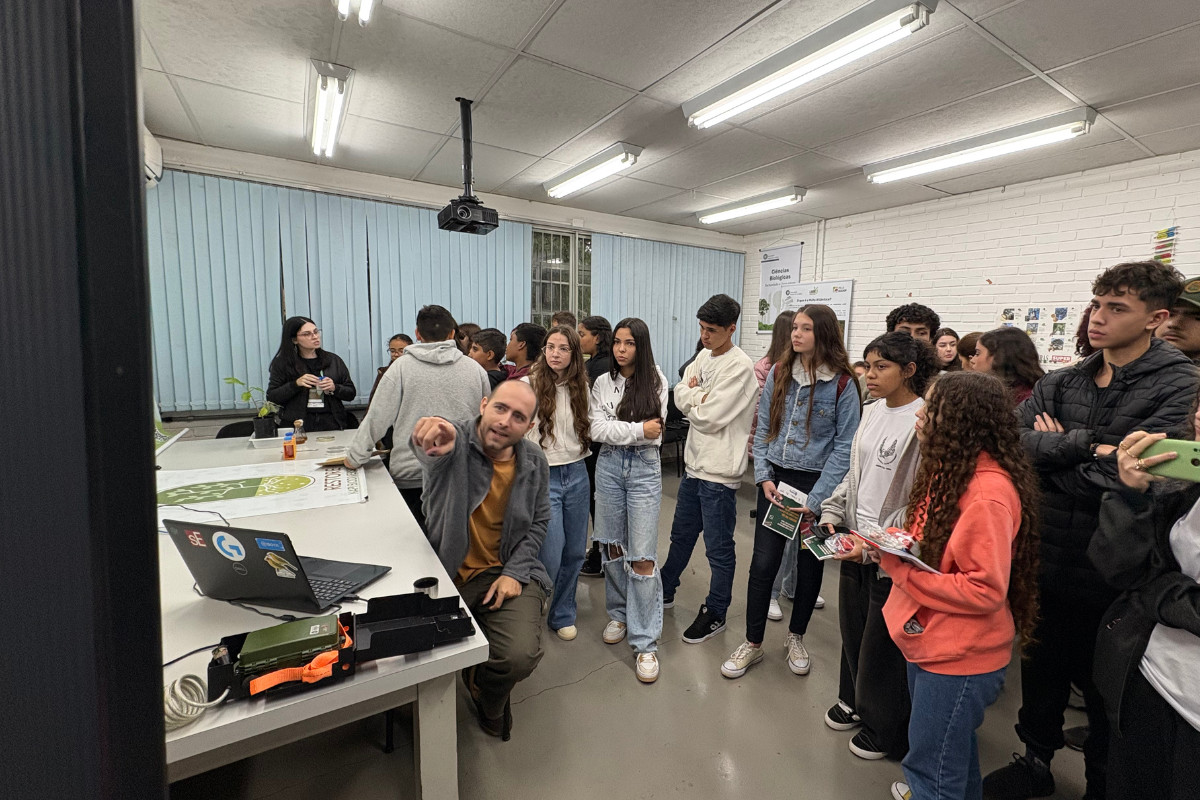 A imagem mostra um pesquisador apontando para um quadro, enquanto alunos prestam a atenção na explicação. O homem está sentado, com os estudantes em pé à sua volta. A turma está em uma sala de laboratório do Centro de Ciências Biológicas da Universidade Estadual de Londrina. Ao fundo, é possível ver uma janela grande com cortinas. As paredes são de tijolos e o espaço é iluminado artificialmente.