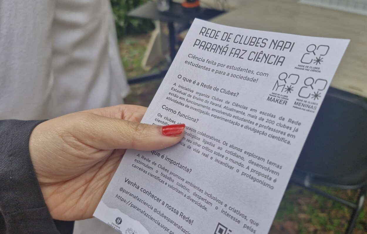 A imagem mostra uma mulher segurando um dos panfletos que foram distribuídos pela equipe NAPI UEL durante a Feira das Profissões. O material de divulgação indica os objetivos da Rede de Clubes e seu papel na sociedade, além de conter um QR Code para que os visitantes possam acessar as redes sociais dos clubes de ciencias da região.