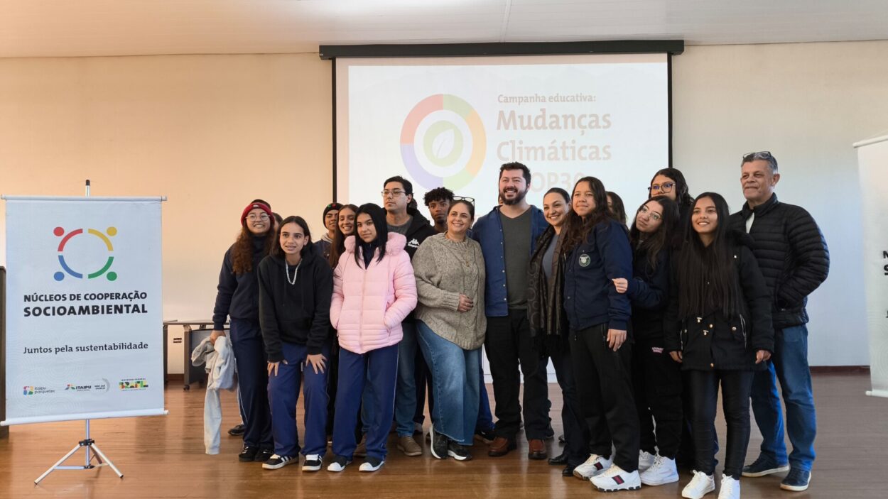 A imagem mostra um grupo de pessoas reunidas em um ambiente interno, um auditório. O grupo é composto por estudantes e alguns adultos (professores e o palestrante). Todos parecem estar felizes e engajados, posando para uma foto em grupo. Ao fundo, há uma projeção com o título da campanha educativa: "Mudanças Climáticas – COP30"**, o que indica que o evento está relacionado a conscientização sobre mudanças climáticas e preparação para a COP30. À esquerda, há um banner com o logotipo dos "Núcleos de Cooperação Socioambiental", e o slogan "Juntos pela sustentabilidade". Também aparecem os logotipos da Itaipu Binacional, Itaipu Parquetec, Ministério do Meio Ambiente e Governo Federal do Brasil, e sinaliza que essa atividade faz parte de um programa oficial de educação ambiental. As roupas das pessoas indicam que o clima está frio, com vários usando casacos, gorros e jaquetas. O ambiente é acolhedor e a atmosfera da imagem transmite união e engajamento com a causa ambiental.