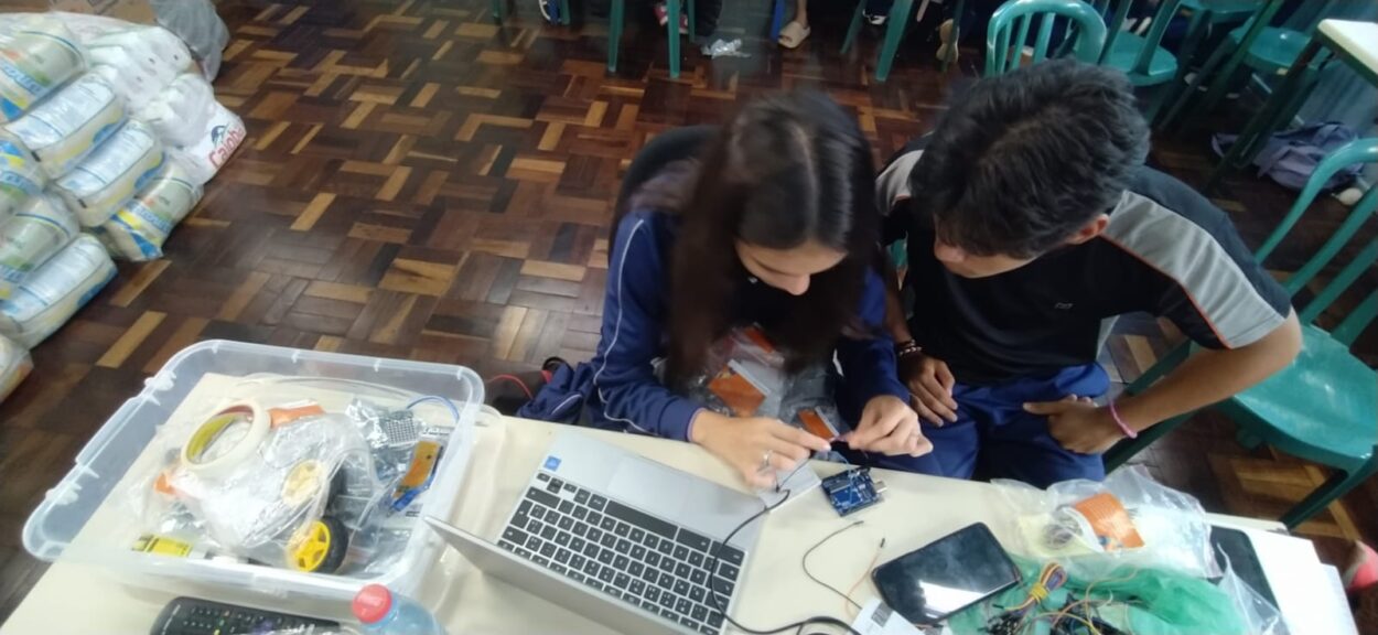 Descrição de imagem: A imagem mostra dois jovens estudantes envolvidos em uma atividade prática de eletrônica ou robótica. Eles estão sentados lado a lado, concentrados em montar ou programar uma placa eletrônica, que está conectada a fios e sensores. Sobre a mesa há uma variedade de materiais, incluindo: um notebook aberto; uma caixa plástica com rodas, fitas adesivas, componentes eletrônicos e peças diversas de robótica; um celular e sacos com fios e pequenos componentes. Ao fundo, é possível ver várias cadeiras verdes ocupadas por outros estudantes. A cena transmite um clima de aprendizado colaborativo e incentivo à tecnologia e inovação