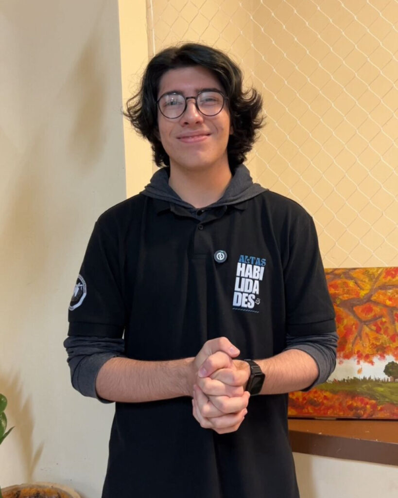A foto é do aluno Davi Angelo Rezende, do Clube Cienteens, no Colégio Instituto de Educação Estadual de Maringá. A imagem mostra um jovem de cabelos escuros e ondulados, usando óculos redondos e uma camisa preta de manga longa com capuz cinza por baixo. Ele está sorrindo levemente e com as mãos unidas à frente do corpo, em um gesto simpático e acolhedor. A camisa tem estampado o texto "ALTAS HABILIDADES" no lado esquerdo do peito. No braço direito da camisa há também um emblema. Ao fundo, vê-se uma parede clara com uma rede protetora e parte de uma pintura colorida com tons de laranja e amarelo. Há também um vaso com planta no canto inferior esquerdo da imagem. A iluminação é suave e natural.
