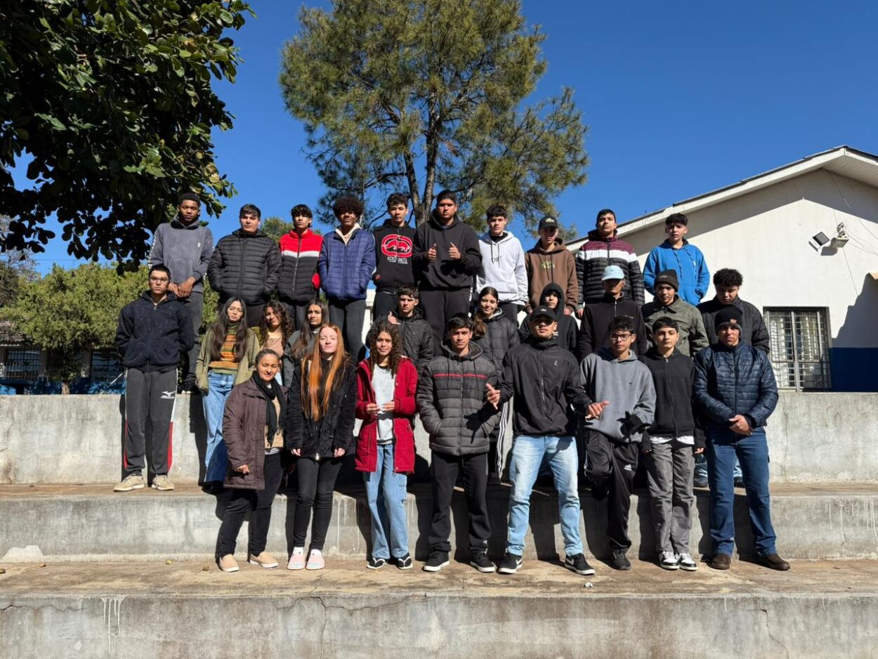 A foto é do Clube Compostagem e Horta Escolar do Colégio Estadual Arthur de Azevedo de São João do Ivaí. A imagem mostra um grupo grande de estudantes e uma adulta que é a professora, posando para uma foto em uma arquibancada de concreto ao ar livre. Eles estão distribuídos em três níveis da arquibancada. A maioria veste roupas de frio, como casacos, jaquetas, toucas e moletons, indicando que o clima está frio. O céu está completamente azul, sem nuvens, e há árvores ao fundo, além de parte de um prédio escolar com janelas. A expressão das pessoas varia entre sorrisos discretos e olhares sérios voltados para a câmera. A cena transmite um ambiente escolar e coletivo.