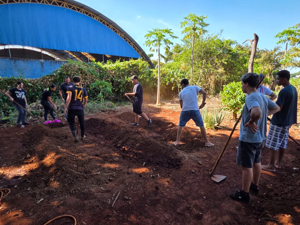 A imagem mostra um grupo de pessoas, em sua maioria jovens, trabalhando em um terreno de terra avermelhada ao ar livre. Ao fundo, há vegetação densa e árvores, como pés de mamão, e também o ginásio esportivo. No centro da imagem, os participantes estão distribuídos ao redor de canteiros elevados de terra, utilizando ferramentas como enxadas e pás para cavar, nivelar e preparar o solo. Todos parecem engajados na atividade. O ambiente é ensolarado e transmite uma sensação de colaboração e trabalho em equipe.